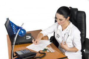 doctor sat at desk writing notes in front of a computer and phone 