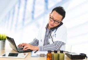 Doctor typing on computer on phone at desk with tablet and bottles on